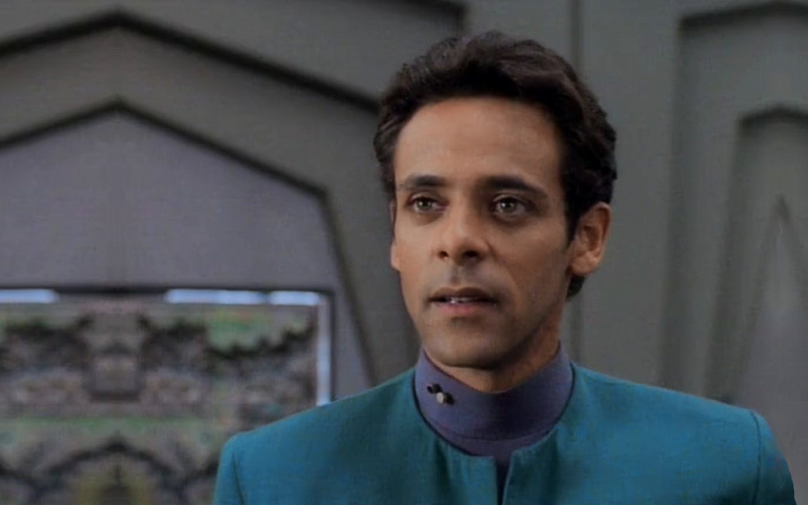 Alexander Siddig, a man with tan skin, hazel eyes, and short, slightly wavy brown hair, as Dr. Julian Bashir. He wears a blue medical uniform and is in front of the geometrically complicated backdrop of the infirmary.