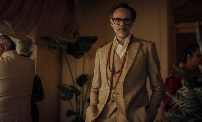 Alexander Siddig, an Arab man with short, dark brown hair, black-rimmed glasses, and white beard and mustache, as Khader Khan. He wears a tan 3-piece suit with two long red-beaded necklaces and a white collared shirt. He stands in a crowded, tan room.
